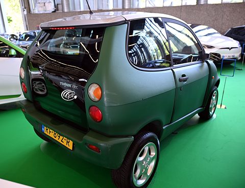 Rear three-quarter of a matt green tall hatchback with dark plastic lower panels and a Dutch plate on green carpet. Opens a larger view