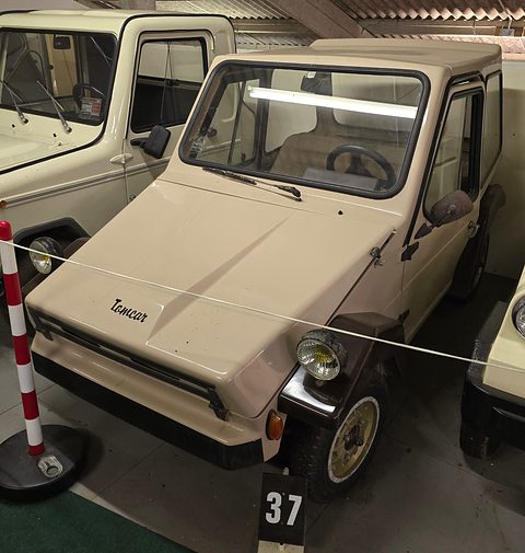 Cream angular four-wheeled microcar with Tomcar script on its bonnet, behind a rope with a number board in front. Opens a larger view