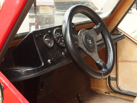 Black three-spoke steering wheel with a badge in its hub, a speedometer and a second dial, seen through the open side. Opens a larger view