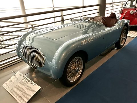 Pale blue open two-seater with a slatted grille and wire wheels beside a printed information panel. Opens a larger view