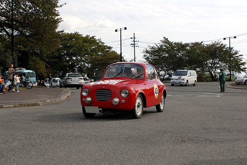Small red coupé with a rally number driving across a car park, trees and spectators behind. Opens a larger view