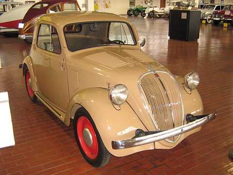 Cream small saloon with red wheels and a chrome-barred grille, on a brick museum floor.