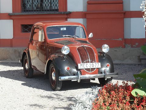 Rust-red and black small saloon parked on gravel in front of a red and white palace wall, flowers in front. Opens a larger view