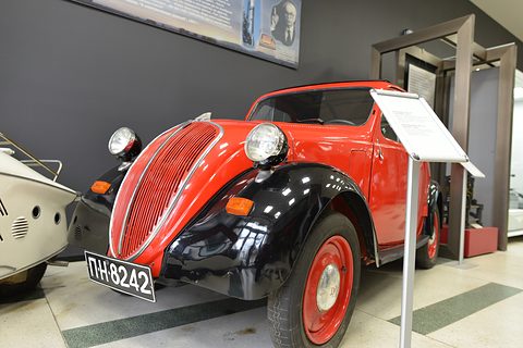 Red small saloon with black wings, red wheels and headlamps on the wings, a museum card on a stand beside it. Opens a larger view