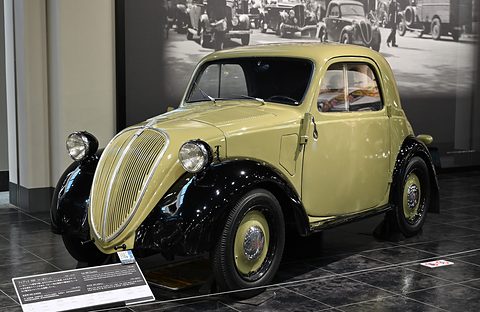 Pale yellow small saloon with black wings, wing-mounted headlamps and a tall grille on a dark tiled floor. Opens a larger view