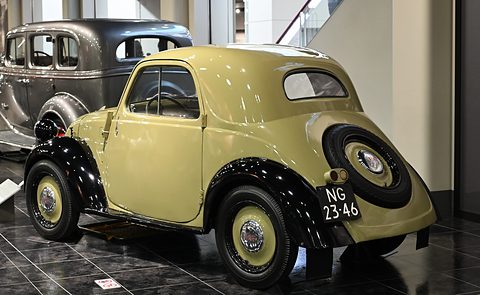 Pale yellow small saloon from the left rear, a spare wheel on its rounded tail and black wings. Opens a larger view