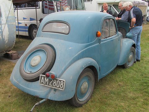Faded blue small saloon from the left rear with a spare wheel on the tail, men looking at it on grass. Opens a larger view