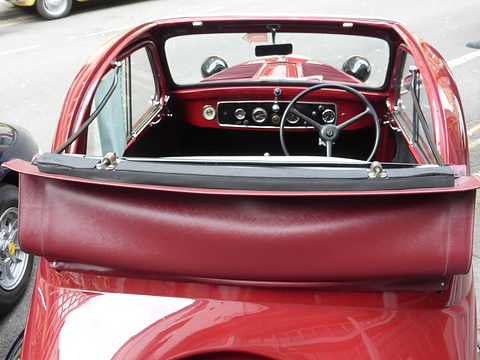 Looking into a small car over a rolled fabric roof: red dashboard with dials, steering wheel and windscreen. Opens a larger view