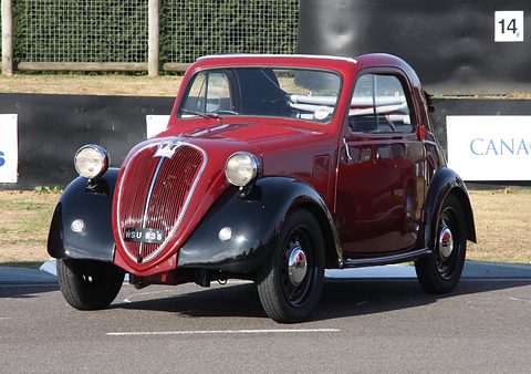 Maroon small saloon with black wings and wing-mounted headlamps, seen from the front on tarmac. Opens a larger view