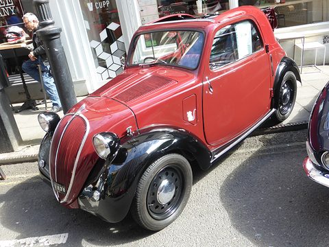 Red small saloon with black wings and wing-mounted headlamps parked in a city street. Opens a larger view
