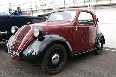 Maroon small saloon with black wings, plate WSU 838, parked on tarmac beside a black open car. Opens a larger view