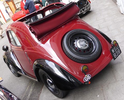Red small car seen from above the rear, its fabric roof rolled back over the tail and a spare wheel on the back.