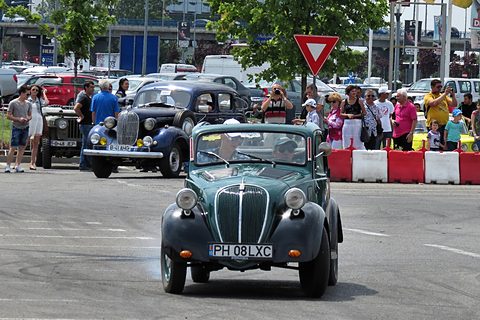 Green small saloon with black wings driving towards the camera, spectators behind barriers. Opens a larger view