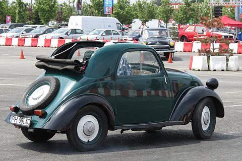 Green small car from the left rear, its fabric roof rolled back and a spare wheel on the tail. Opens a larger view