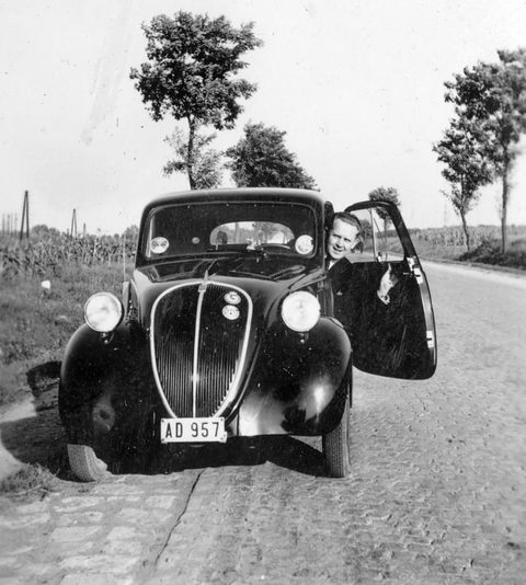 Black and white: a small dark saloon on a cobbled road, a man leaning out of the open door.