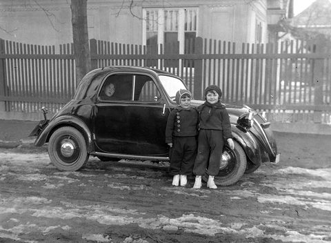Black and white: a small dark saloon parked by a fence, two children in winter clothes standing at its side. Opens a larger view