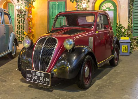 Maroon small saloon with black wings and a sign reading FIAT TOPOLINO 1943 569 cc, a painted street set behind. Opens a larger view