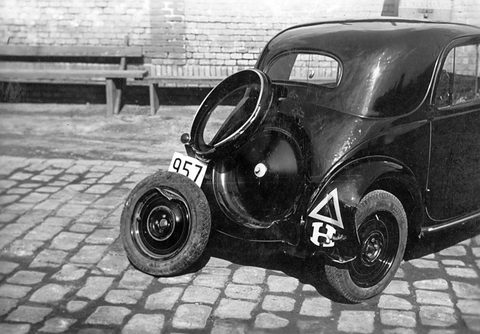 Black and white: a small dark saloon tilted on cobbles with its rear wheel detached beside it. Opens a larger view