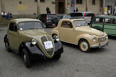 Two small saloons on a square: a dark green and black one with a tall grille, a beige one with a wide grille. Opens a larger view