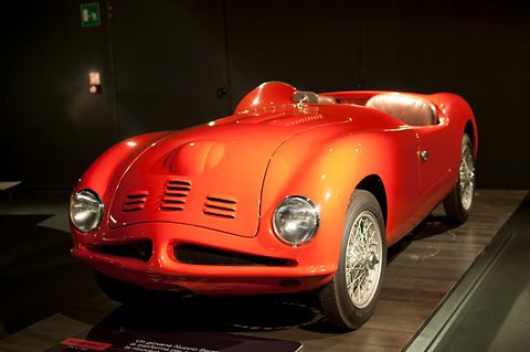 Orange open two-seat sports car with louvres on its nose and wire wheels in a dark gallery. Opens a larger view