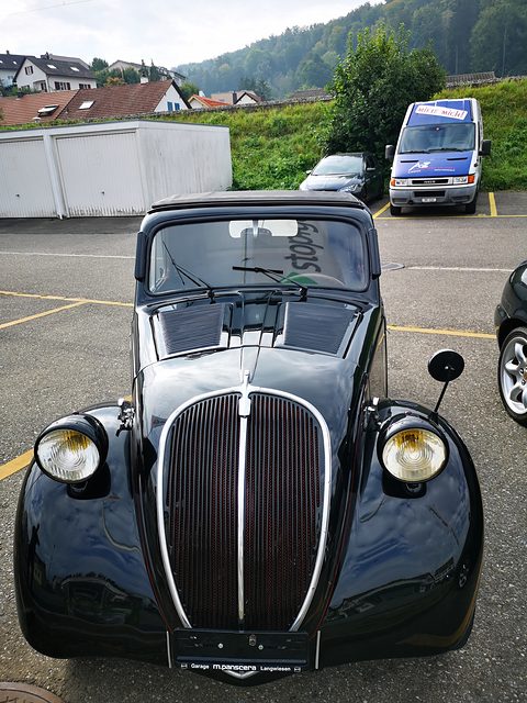 Black small saloon from the front: tall grille, headlamps on the wings, a car park and hillside behind. Opens a larger view