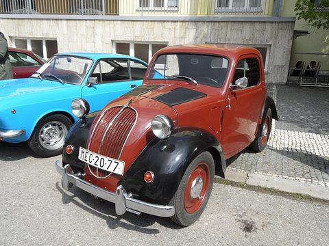 Rust-red small saloon with black wings, plate MEC 20-77, parked on cobbles beside a blue coupé. Opens a larger view