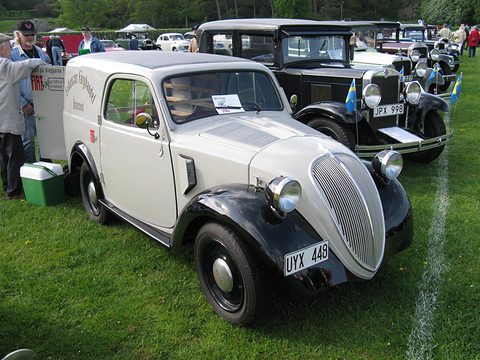 Cream small van with black wings and a tall grille parked on grass with vintage cars behind. Opens a larger view