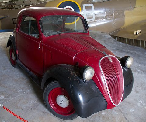 Maroon small saloon with black wings and red wheel centres beneath the wing of a camouflaged aircraft. Opens a larger view