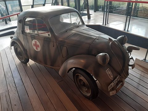 Small saloon in mottled camouflage paint with a red cross on its door, on a wooden display floor. Opens a larger view