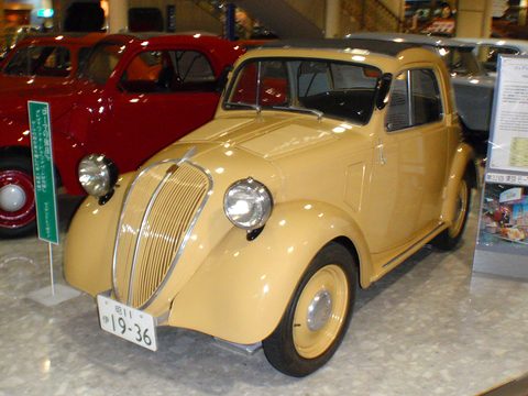 Cream small saloon with a chrome-barred grille and a Japanese plate in a museum line-up. Opens a larger view