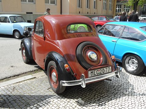Rust-red small saloon from the left rear with a spare wheel on the tail, on cobbles. Opens a larger view