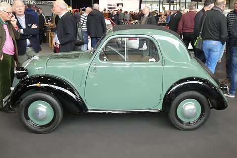 Mint-green small saloon with black wings, seen side-on in a crowded show hall. Opens a larger view