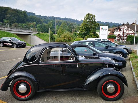 Black small saloon side-on in a car park, red wheels and a short tail. Opens a larger view