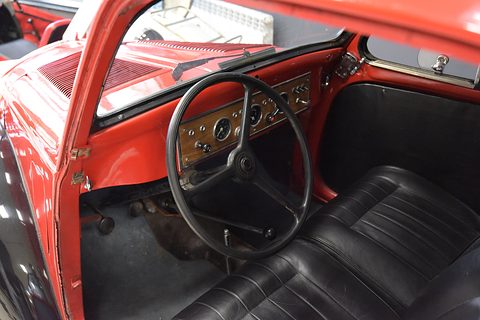 Wood-faced dashboard with small dials, a thin three-spoke steering wheel and black leather seat. Opens a larger view