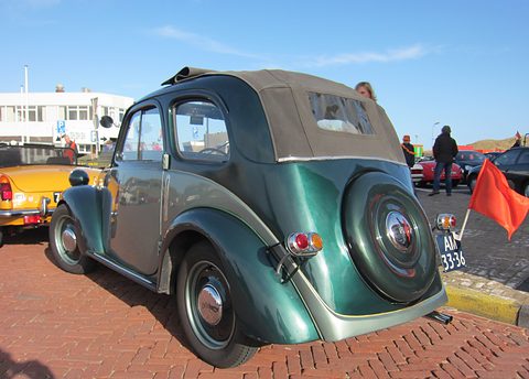 Two-tone green small car from the left rear with its fabric roof closed and a spare wheel on the tail. Opens a larger view