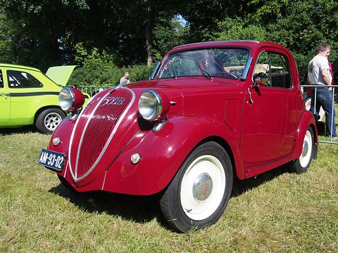 Red small saloon with white wheels and a tall grille parked on grass, a green car behind. Opens a larger view