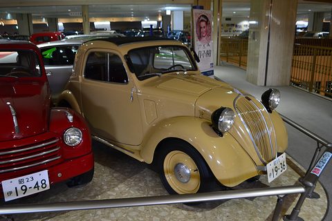Yellow small fastback saloon with headlamps on the wings, behind a rail in a museum hall. Opens a larger view