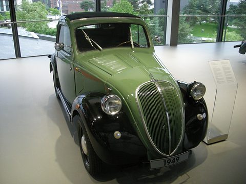 Green small saloon with black wings and a 1949 plate on a white display floor by tall windows. Opens a larger view