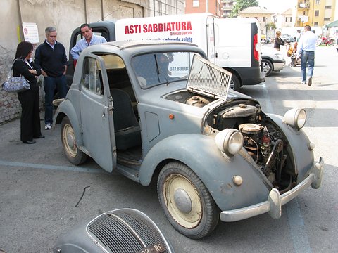 Weathered grey small saloon with its bonnet up and door open, visitors standing behind. Opens a larger view