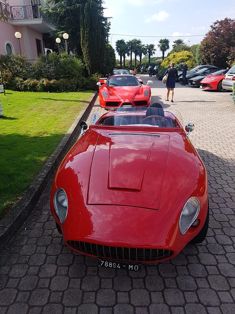 Red open two-seater with a long bonnet scoop, seen from the front on a paved drive with Ferraris behind. Opens a larger view