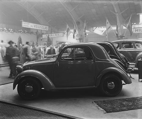 Black and white: a small saloon on a show stand under a high hall roof, visitors beyond. Opens a larger view