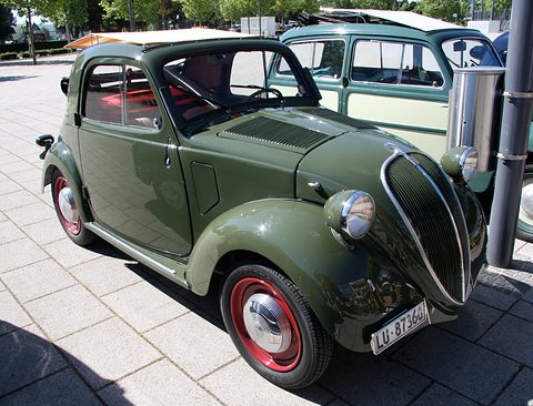 Green small car with red wheels and wing-mounted lamps, its roof open, on paving. Opens a larger view
