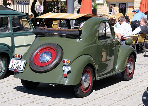 Green small car from the left rear, roof folded open, red wheels and a spare on the tail. Opens a larger view
