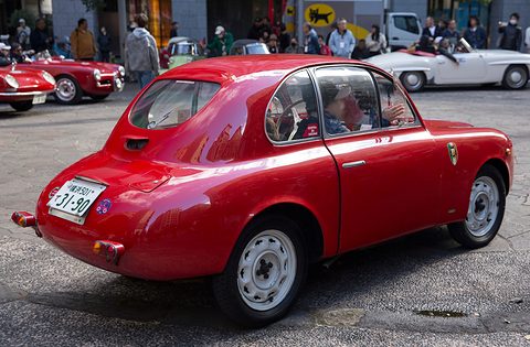 Red small coupé from the right rear with wrap-round windows reaching into the roof. Opens a larger view