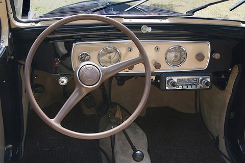 Cream dashboard with two round dials and a radio, a large two-spoke steering wheel in front. Opens a larger view