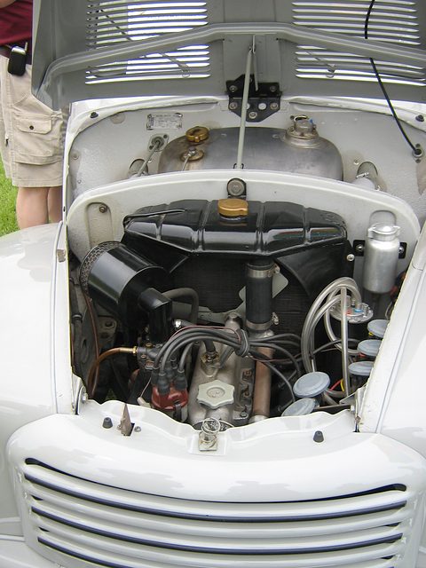 Engine bay under a raised white bonnet: four-cylinder engine with its radiator behind, a slatted grille below. Opens a larger view