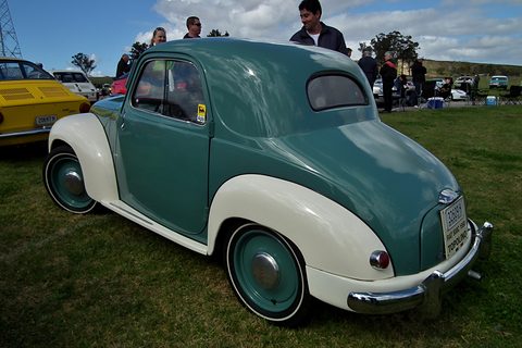 Green and cream small saloon from the left rear on grass, people behind. Opens a larger view