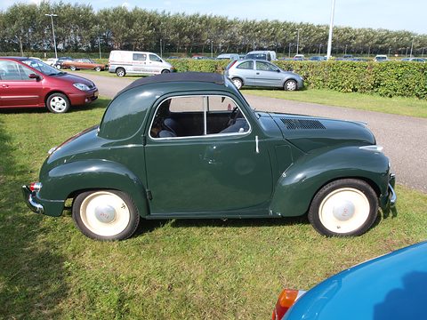 Dark green small saloon side-on on grass with cream wheels, parked cars behind. Opens a larger view