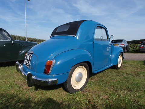 Blue small saloon from the left rear, black fabric roof and cream wheels, under a blue sky. Opens a larger view