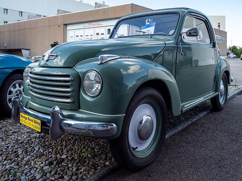 Grey-green small saloon with a horizontal-bar grille and faired-in headlamps on gravel. Opens a larger view